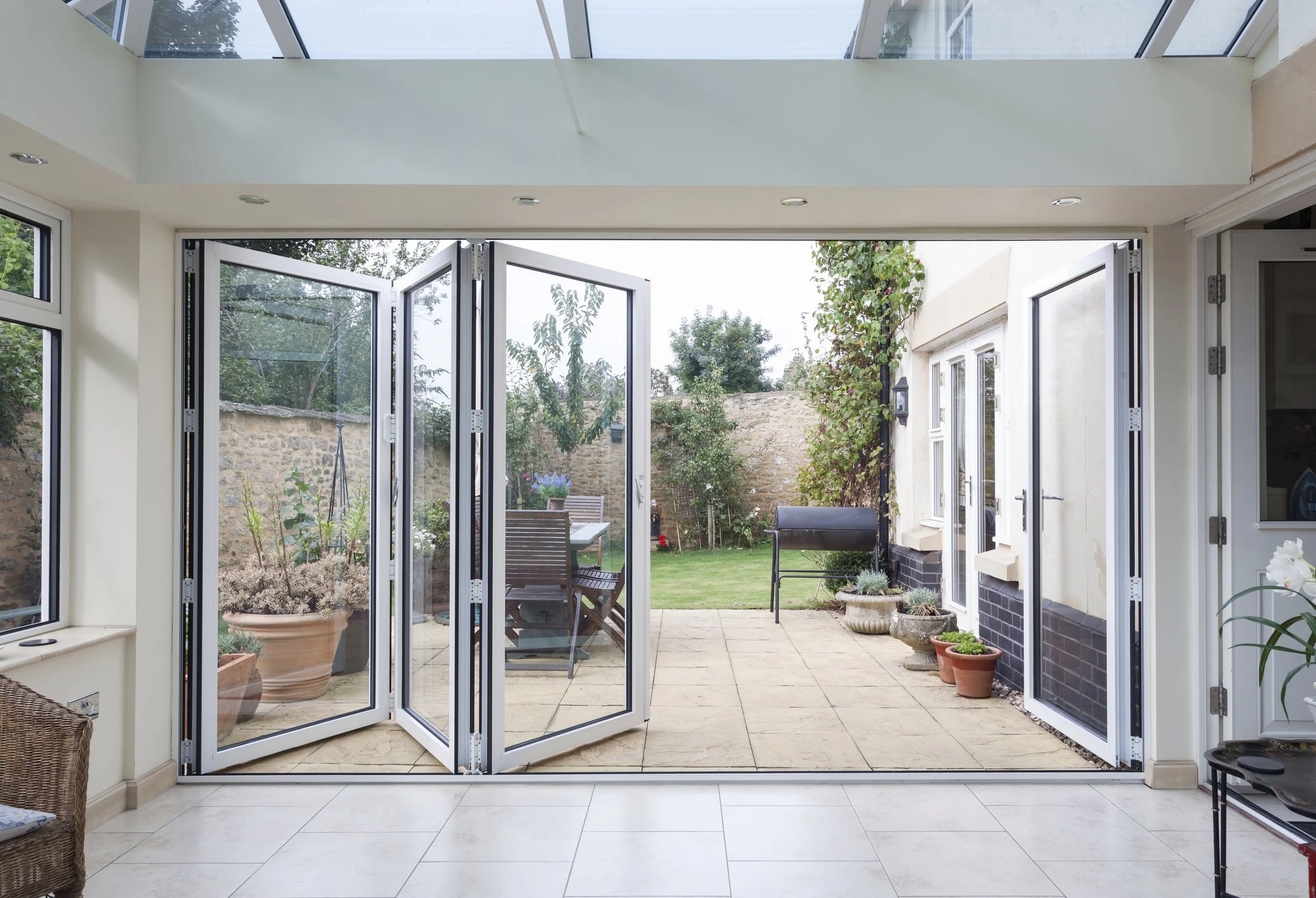 Open glass patio doors leading to a backyard garden with outdoor furniture, potted plants, and a lawn surrounded by a brick wall.
