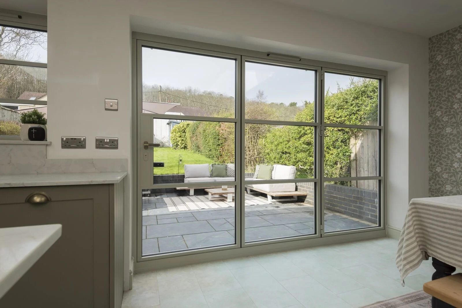 View of an outdoor patio with white cushioned furniture, surrounded by greenery, seen through glass sliding doors from inside a house.