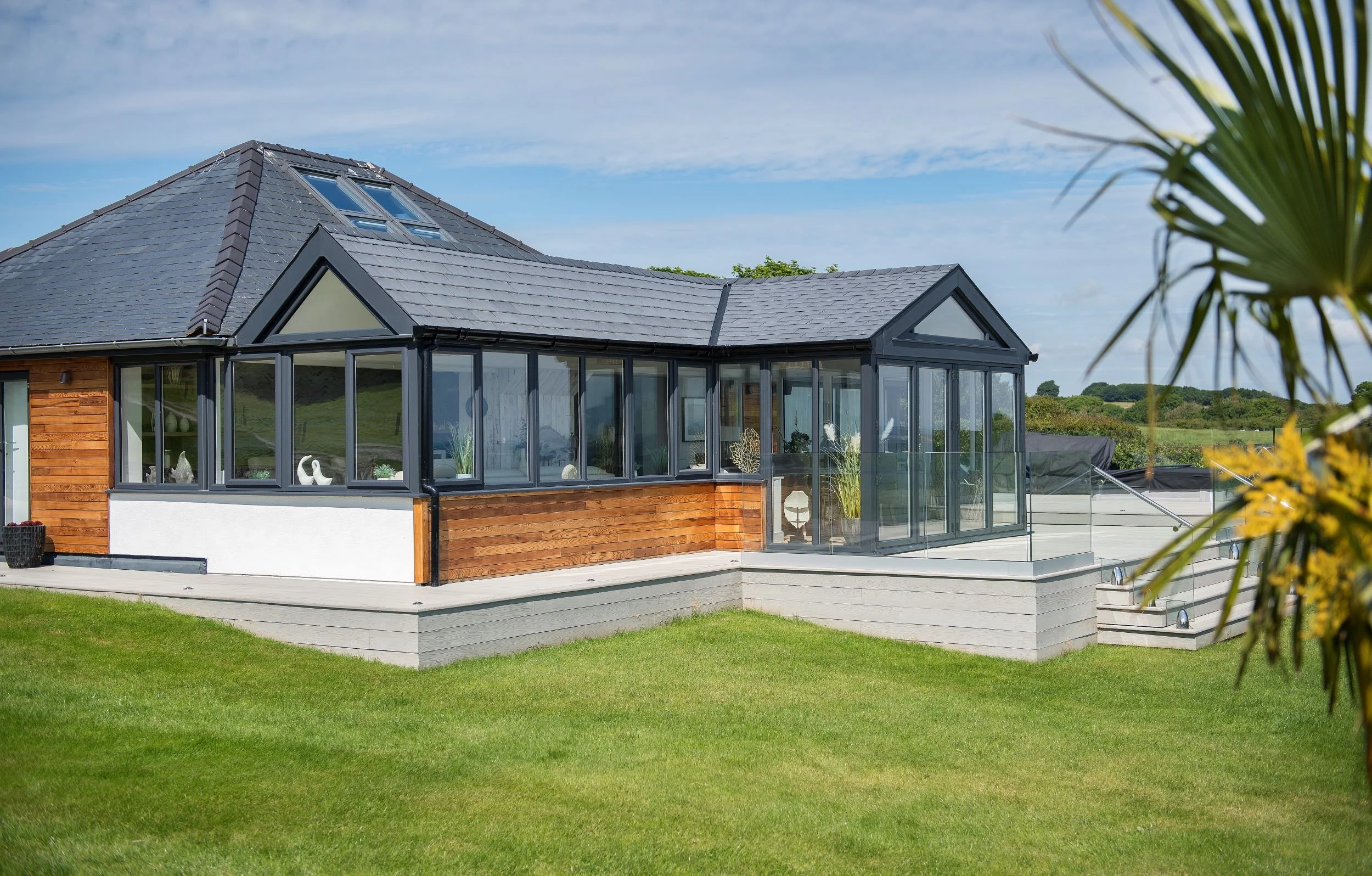 A modern house with a black roof, large glass windows, and a deck with steps, set in a green field under a blue sky.