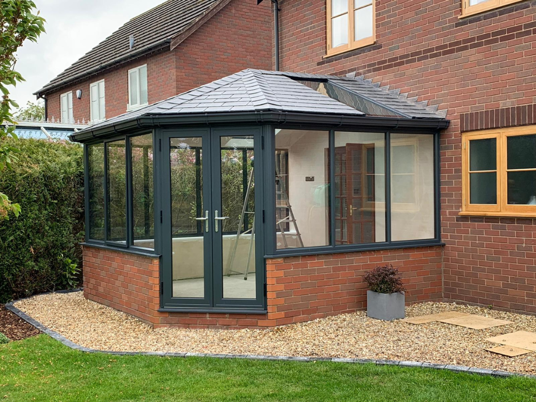 A brick house with a sunroom or conservatory attached to the back, surrounded by a gravel pathway and green lawn, with a shrub in a gray planter near the porch.