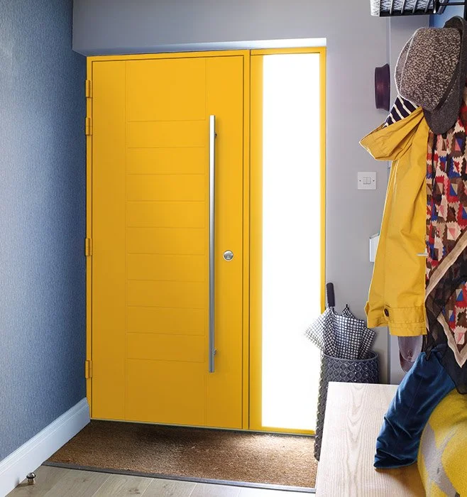 A bright yellow door with a vertical silver handle, next to a lighted white panel, in a modern entryway. Coats, hats, and bags are hanging nearby.