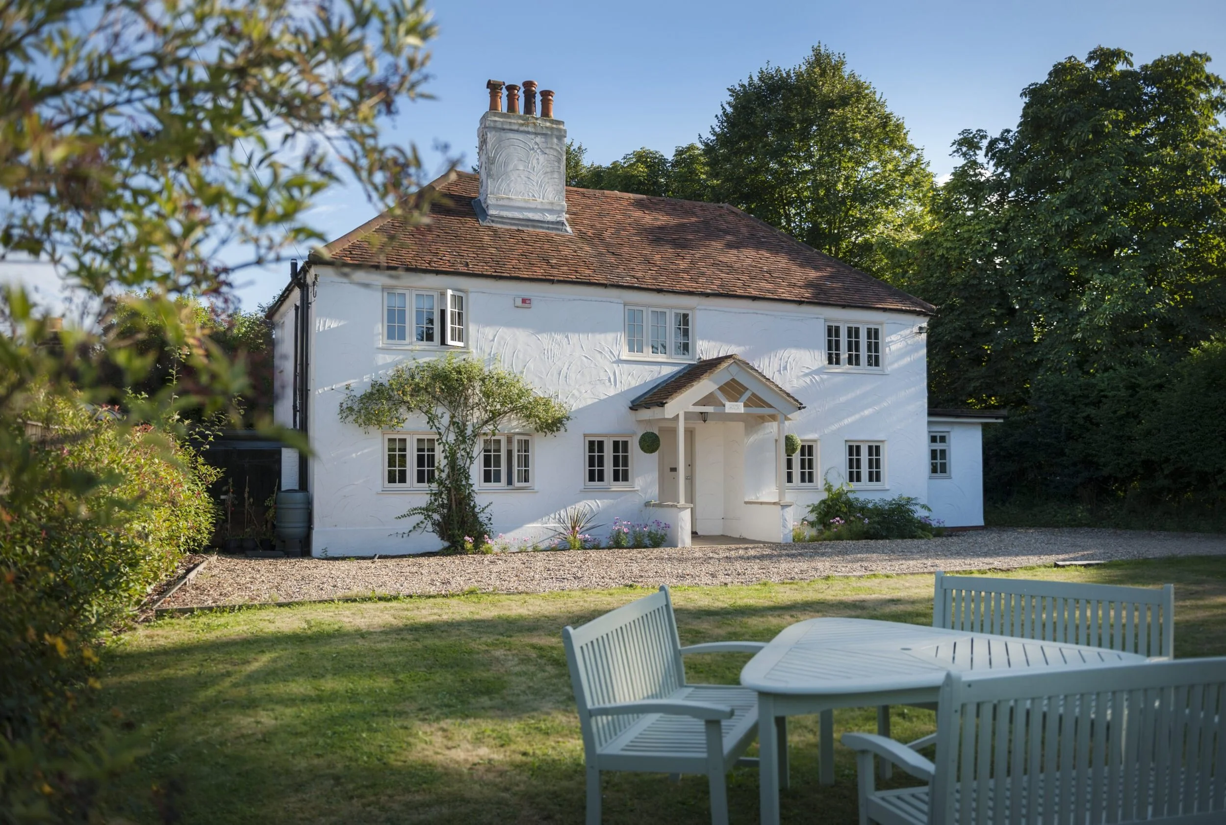 A white two-story house with a red tile roof, surrounded by trees and a lawn, with outdoor furniture in the foreground.