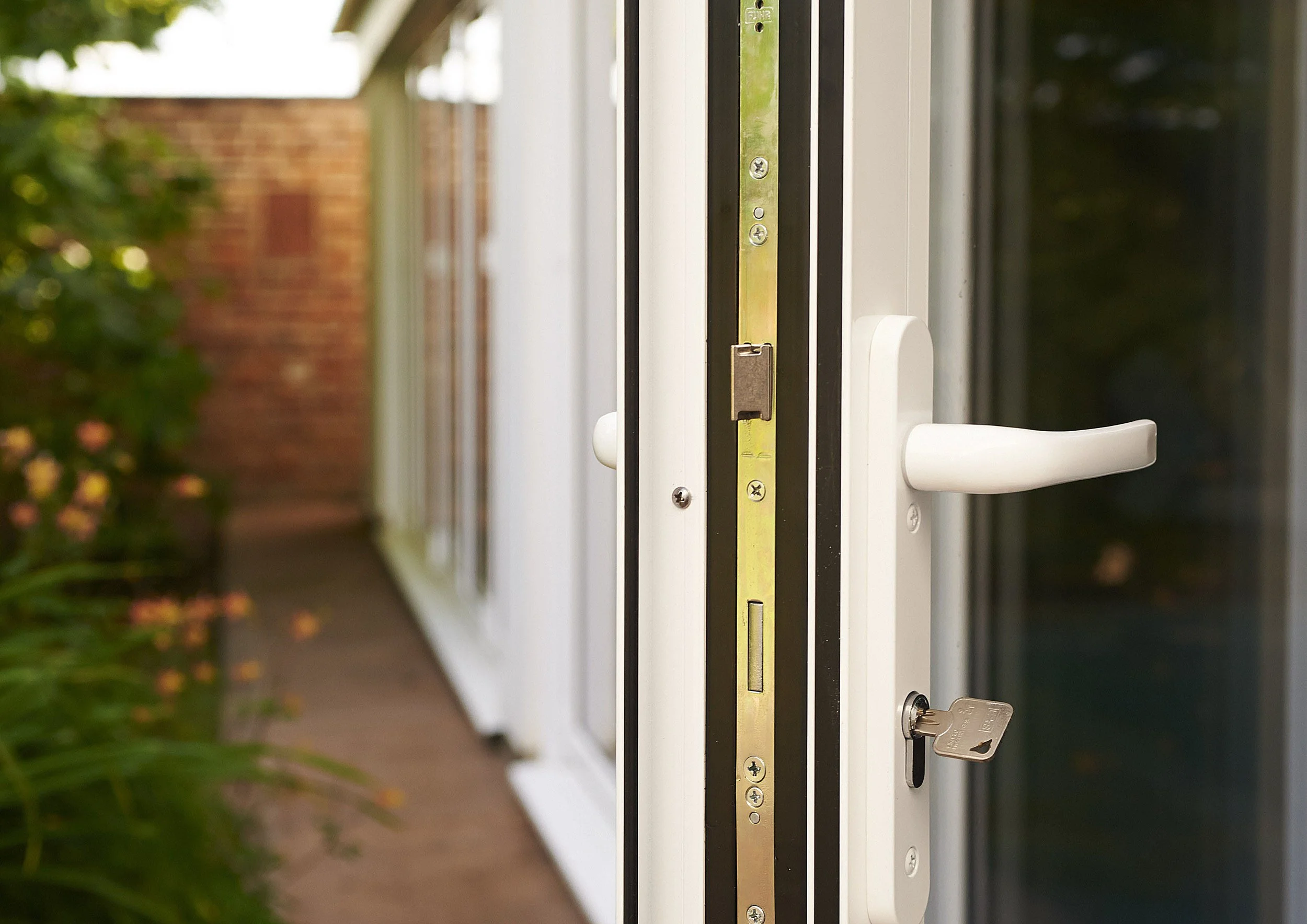 Close-up of a white door handle and lock on a glass door, with a garden and brick wall in the background.