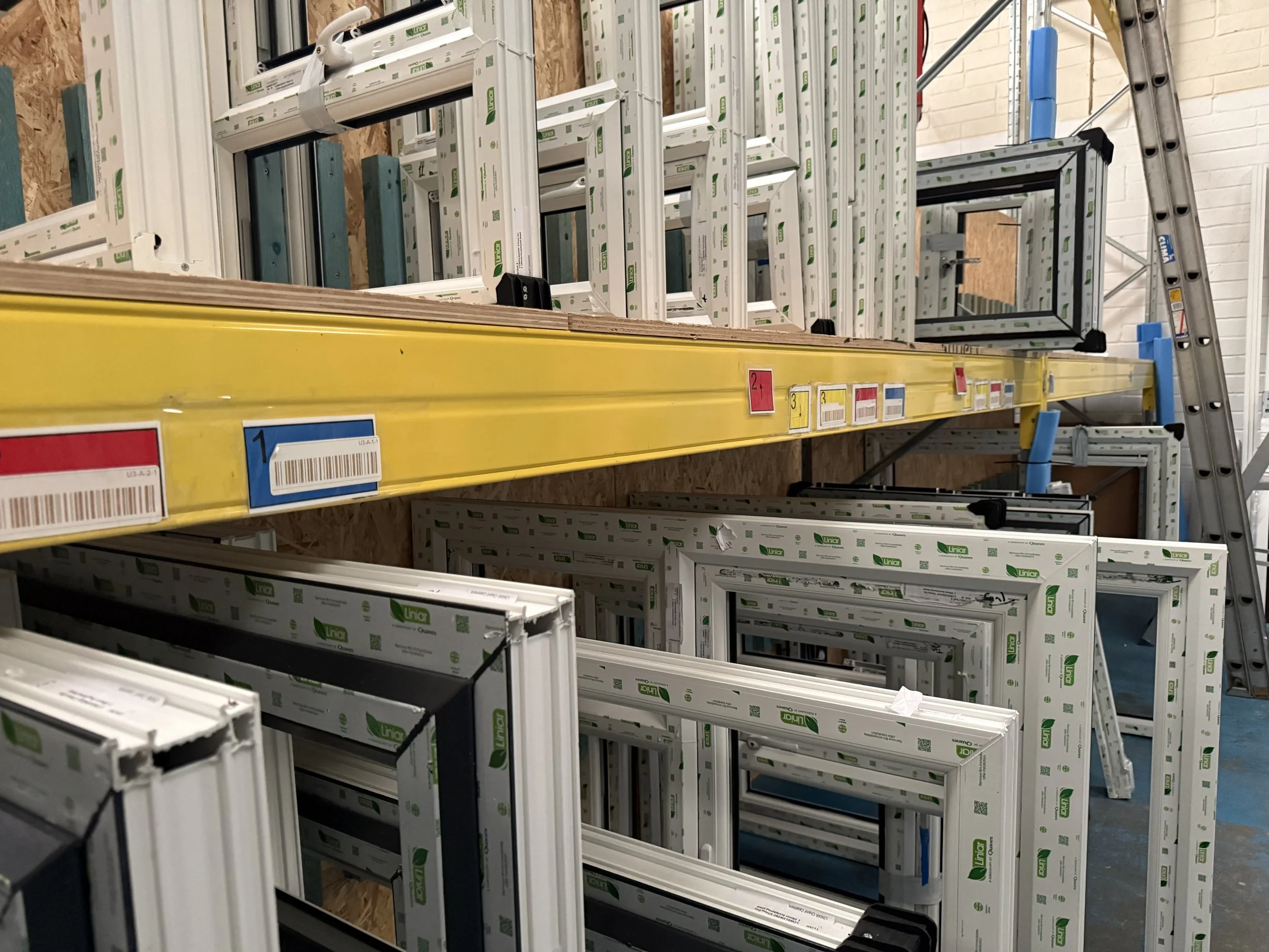 Store aisle with window frames stacked on shelves and a ladder in the background.