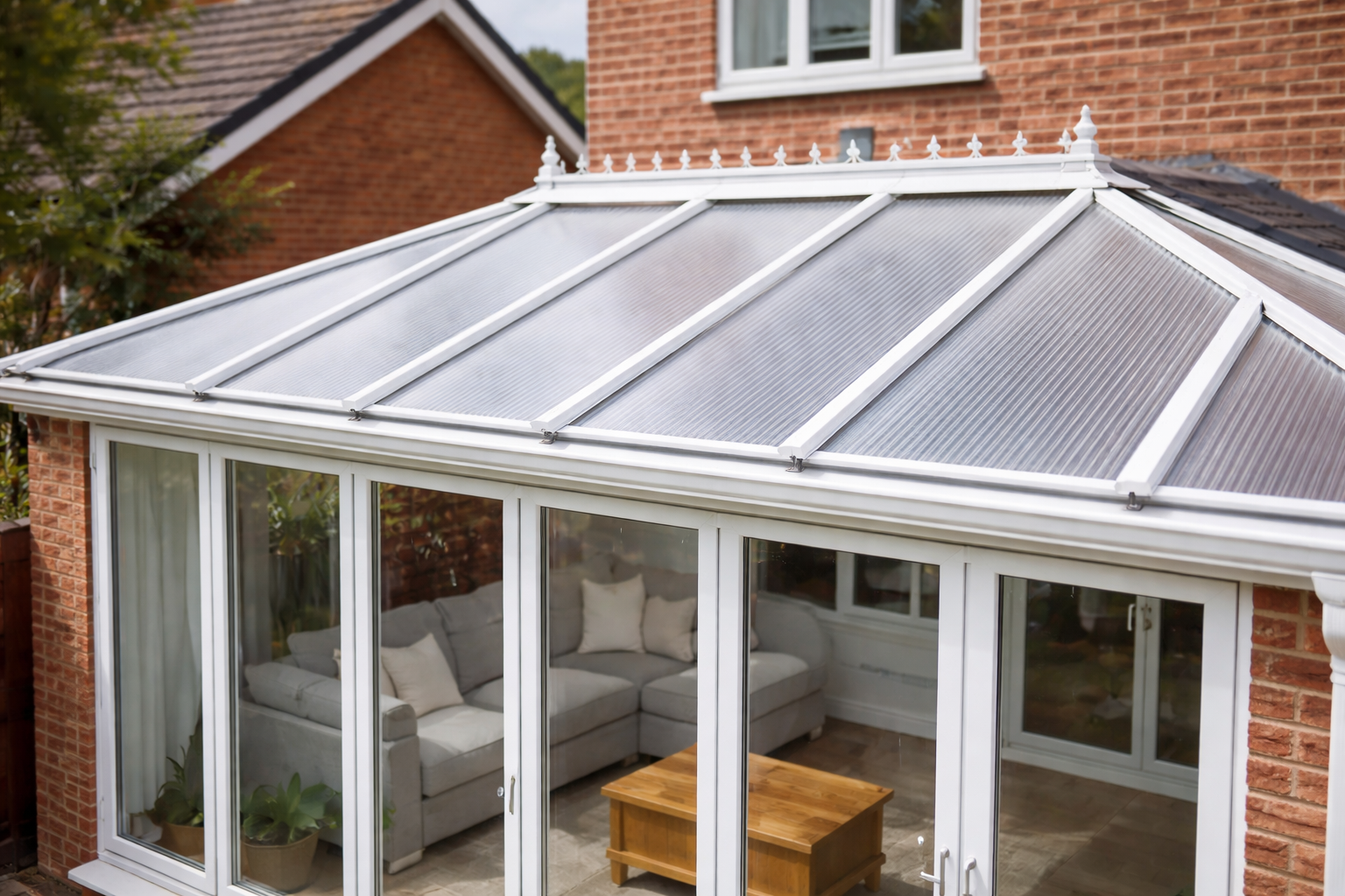 A glass conservatory with a pitched roof attached to a brick house, furnished with a gray sectional sofa, pillows, a small wooden coffee table, and plants inside.