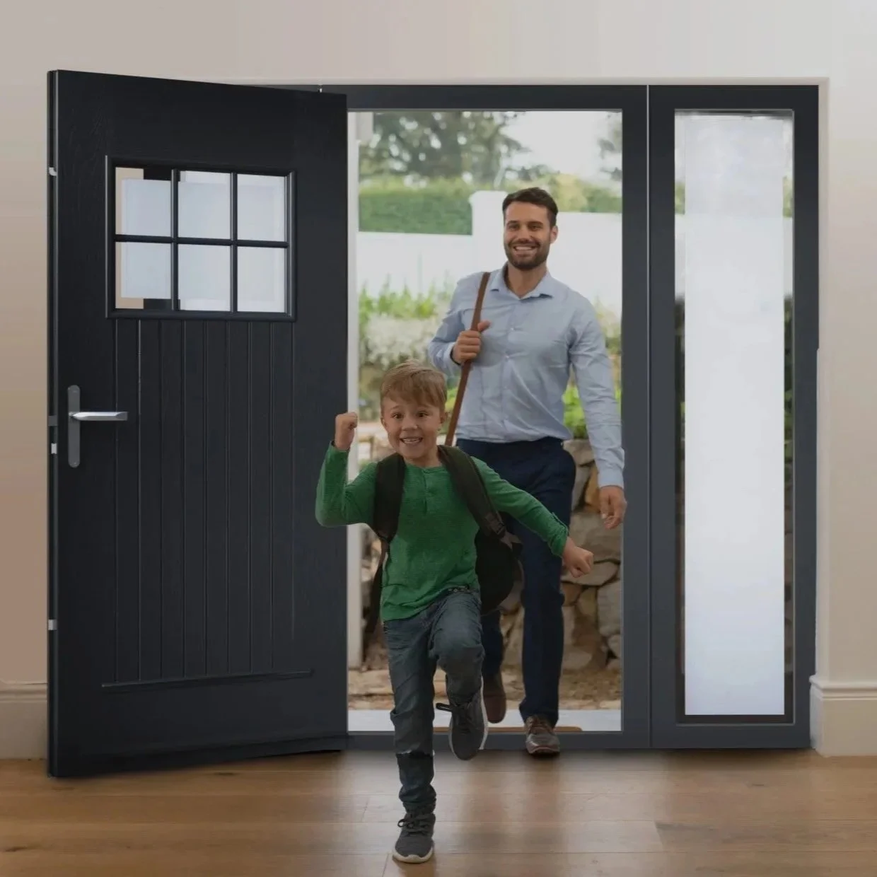 A man and a boy are enthusiastically running indoors toward the camera through a doorway, with the man carrying a shoulder bag and the boy wearing a backpack, suggesting they are arriving home.