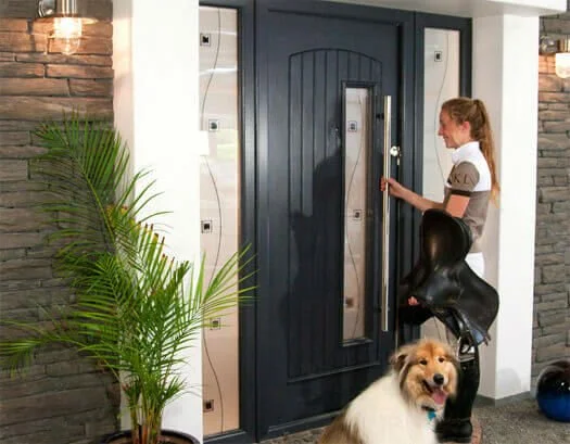 A woman is unlocking a front door with a large black handle, standing next to a dog and a potted plant inside the house, with a brick wall and modern interior design in the background.