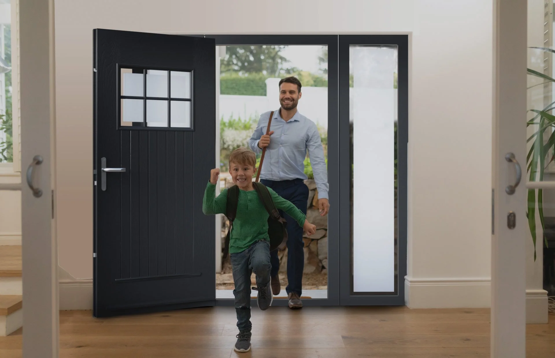 A young boy and an adult man, likely his father, are running joyfully through a door into a house. The boy is wearing a green shirt and backpack, while the man is dressed in a light blue shirt and dark pants. The background shows a porch and greenery