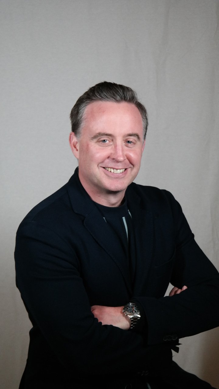Portrait of a smiling man with folded arms, wearing a black jacket and a wristwatch, against a plain gray background.