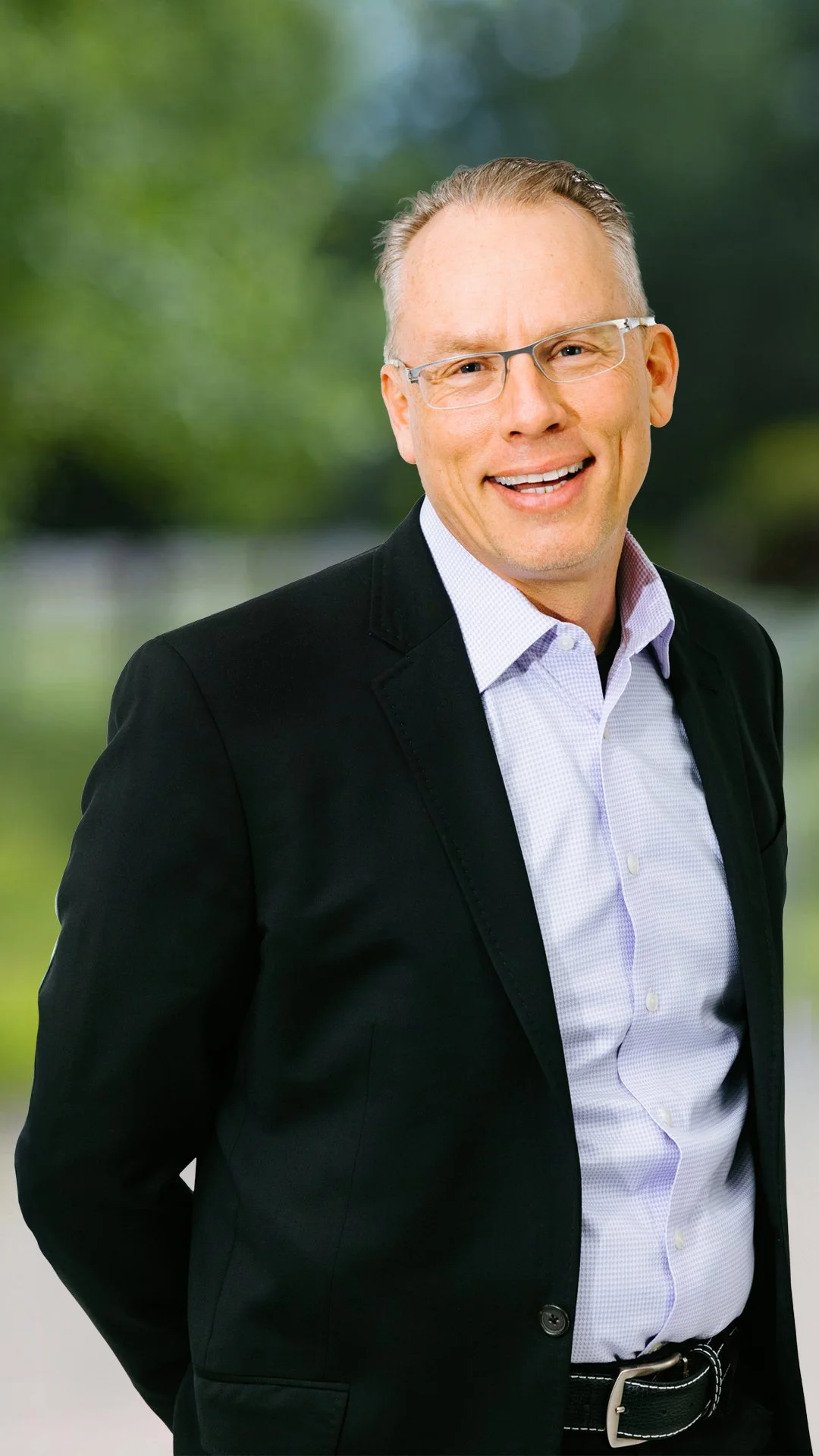A smiling man with glasses, wearing a black jacket and a light purple shirt, standing outdoors with a blurred green background.
