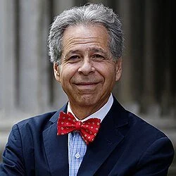 Portrait of an elderly man with gray hair, wearing a navy blazer, blue shirt, and red polka dot bow tie, standing outdoors with a blurred background.