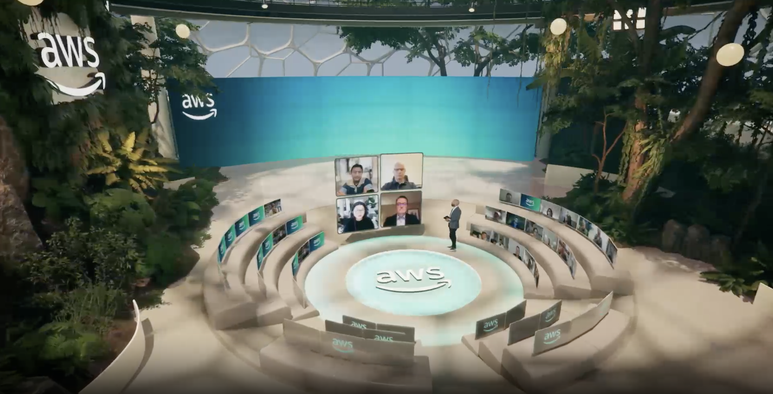 Virtual conference setup with a large circular AWS logo on the floor, multiple screens displaying diverse participants arranged in a semi-circle, and a presenter standing with a tablet. The background features trees and Amazon Web Services branding.