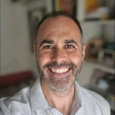 Close-up of smiling man with short dark hair and beard, wearing a light gray collared shirt, in an indoor setting.