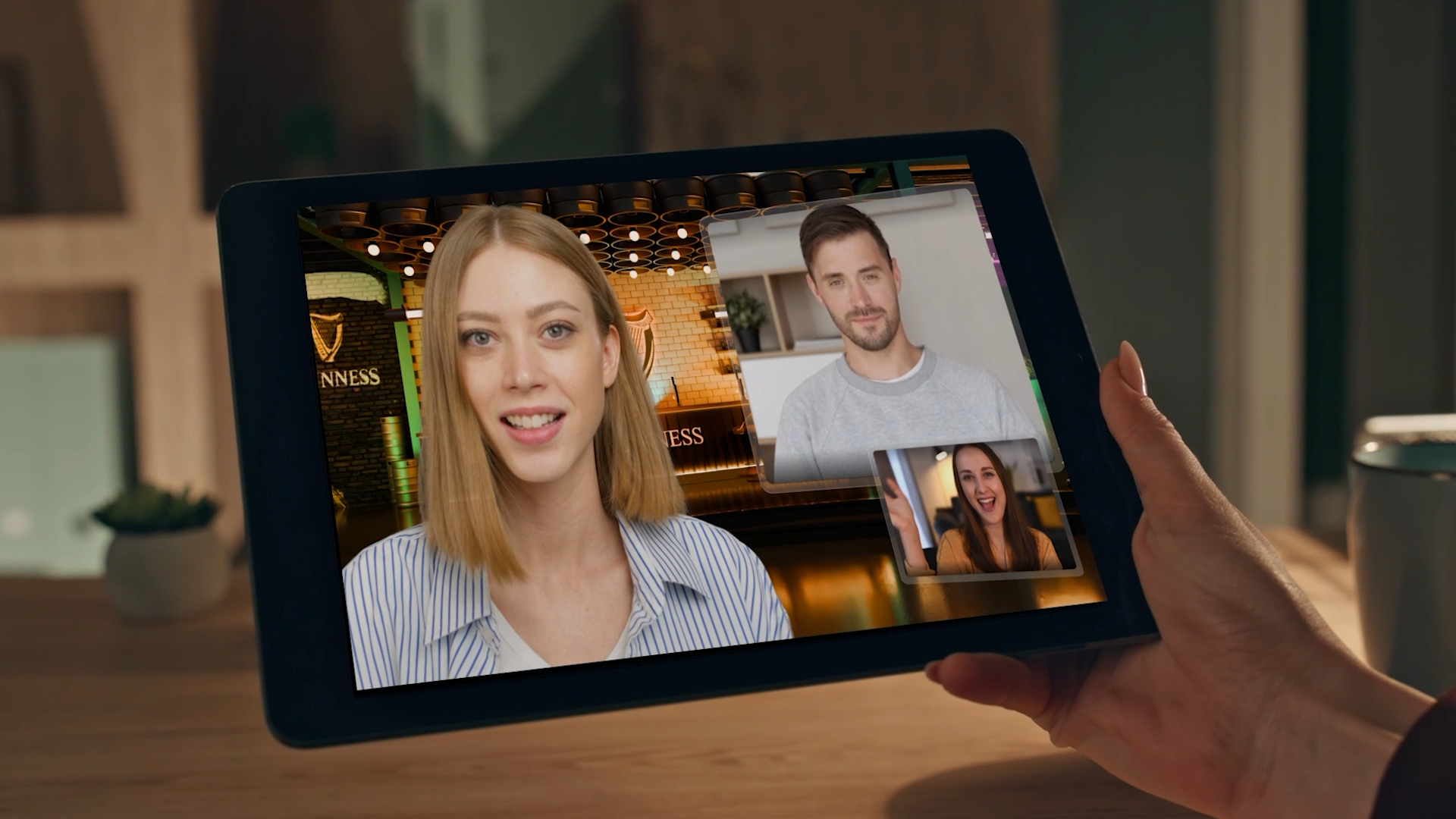 Person holding a tablet with a video call of four women, two joined in larger screens and two in smaller screens, in a room decorated with Guinness signs and bar elements in the background.