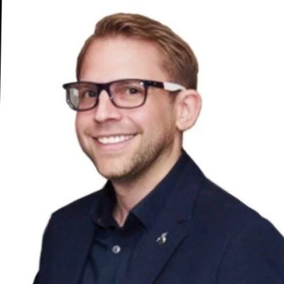 Portrait of a young man with light brown hair, wearing glasses and a dark blazer, smiling against a white background.
