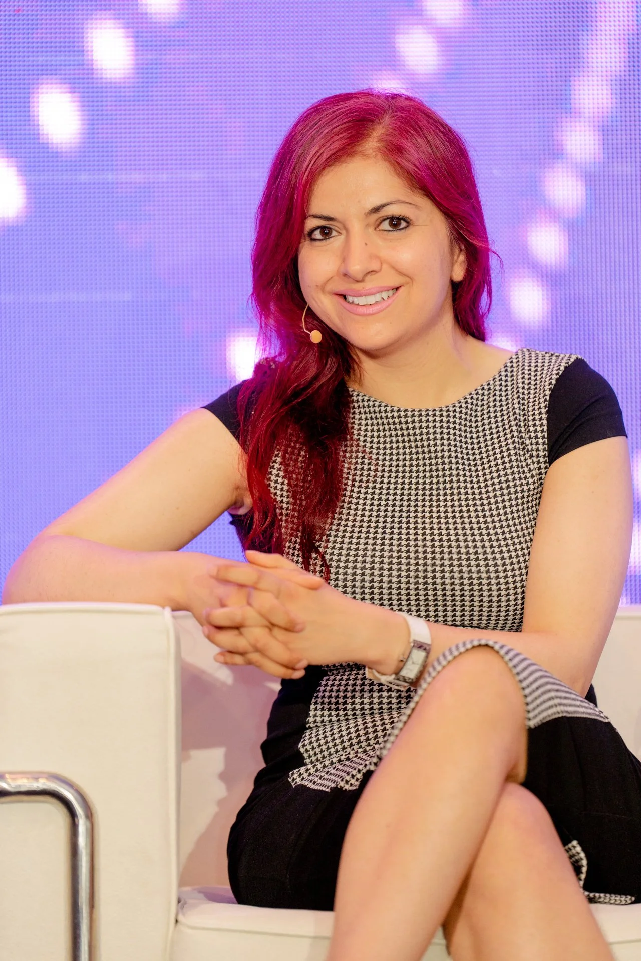 A woman with red hair sitting on a white chair, smiling, against a purple background with bokeh lights.
