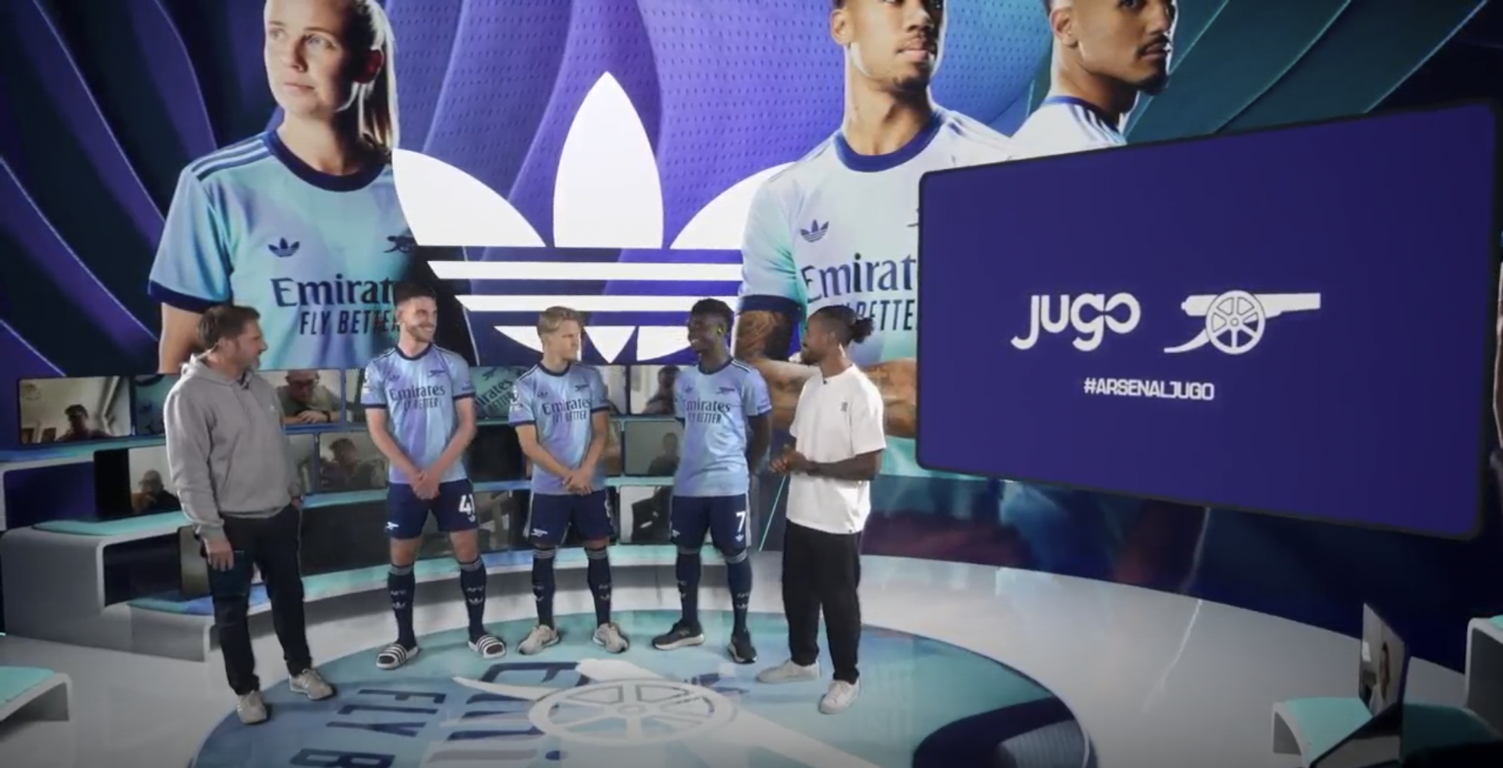 Four men are in a television studio with large screens displaying soccer players and logos. The men are dressed in athletic and casual clothing, standing on a circular platform with an Arsenal logo. The studio features a modern, tech-focused design with multiple monitors and a background of purple and blue shades.