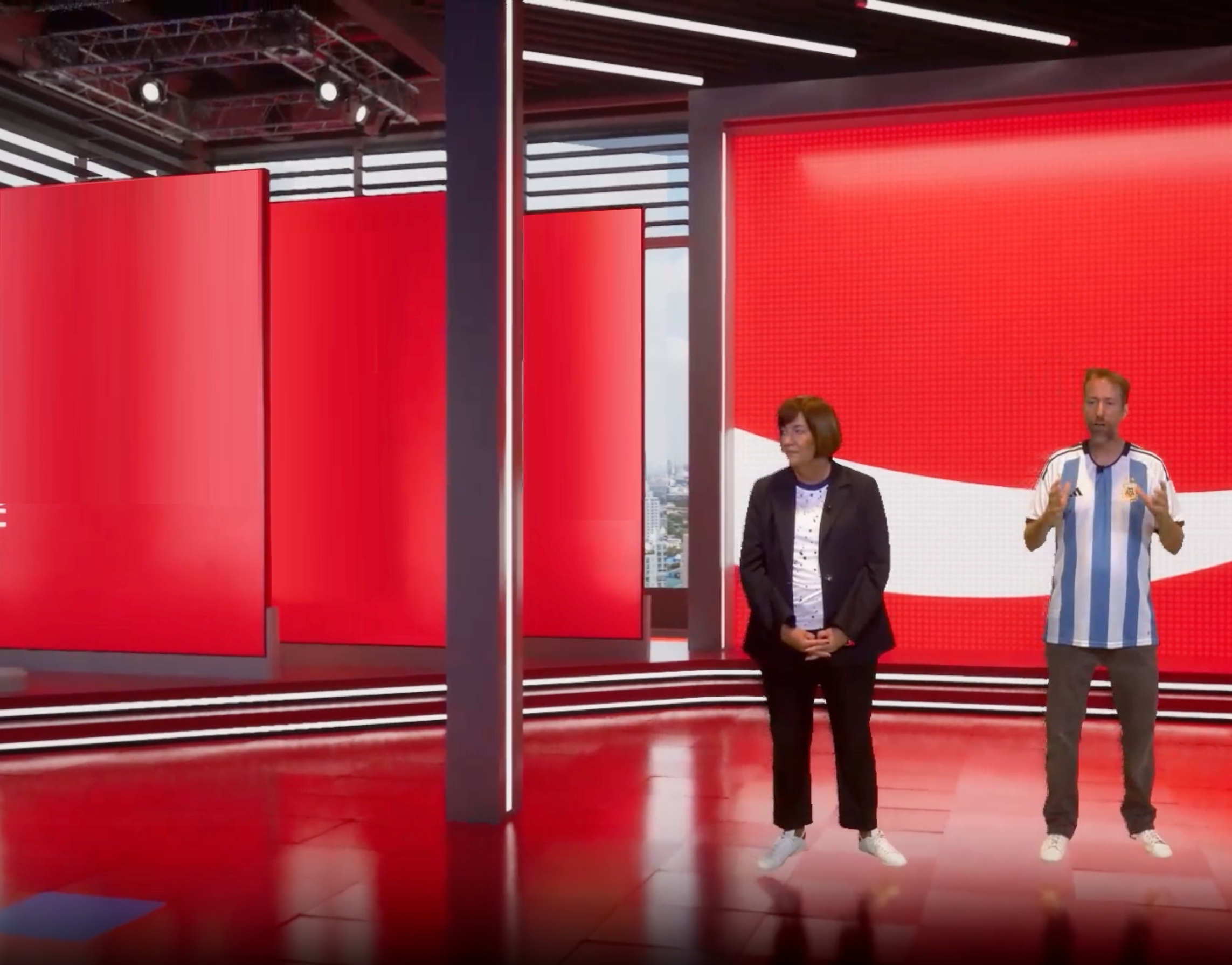Two people standing in a modern studio with a red background, one woman in black blazer and jeans, one man in Argentina soccer jersey.