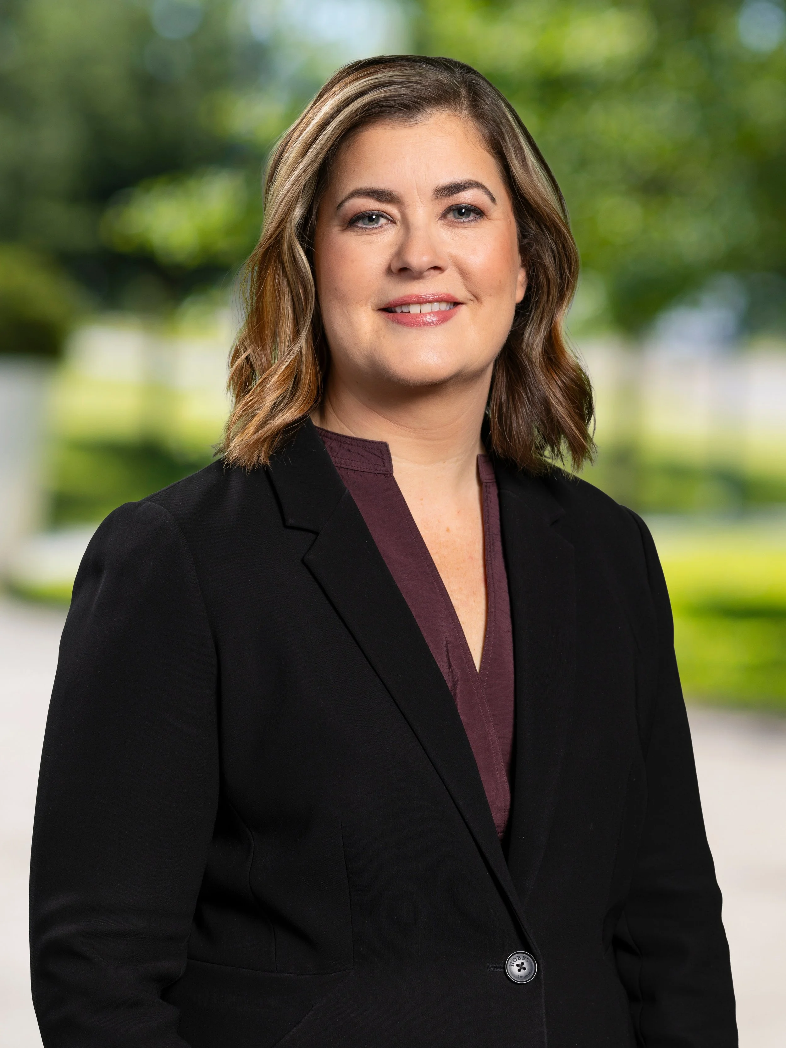 Professional woman with shoulder-length brown hair, wearing a black blazer and a maroon blouse, standing outdoors with blurred green trees in the background.