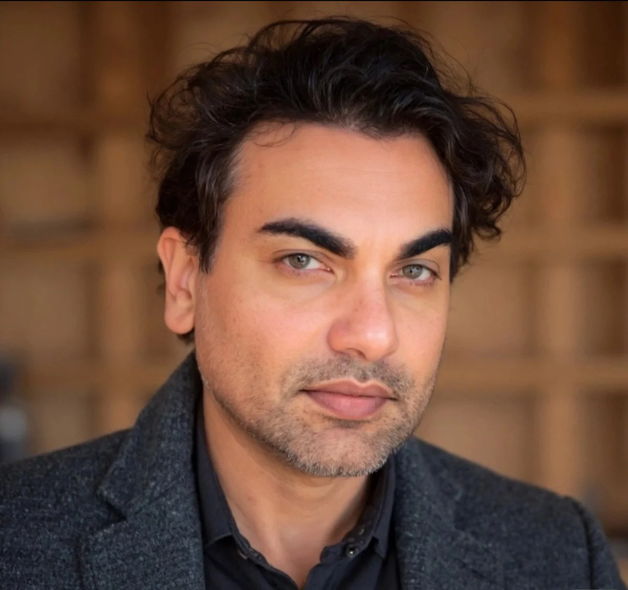 Close-up of a man with dark wavy hair and light-colored eyes, wearing a dark blazer and shirt, against a blurred brownish background.