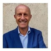 A smiling man with a shaved head, wearing a blue jacket and a light blue shirt, standing against a plain background.