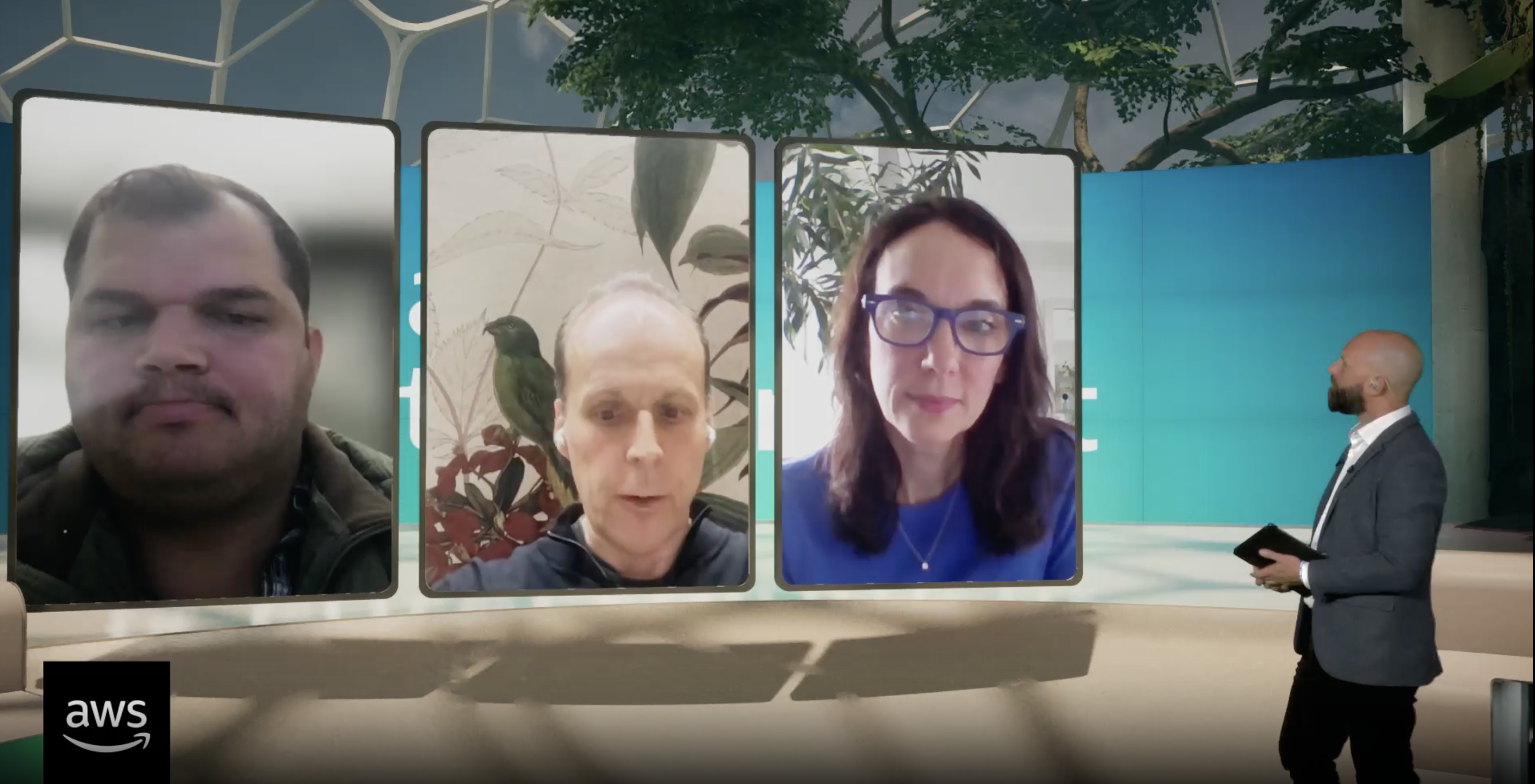 Virtual conference with four participants displayed on screens and a presenter standing on stage, with a nature-themed backdrop and an aws logo in the corner.