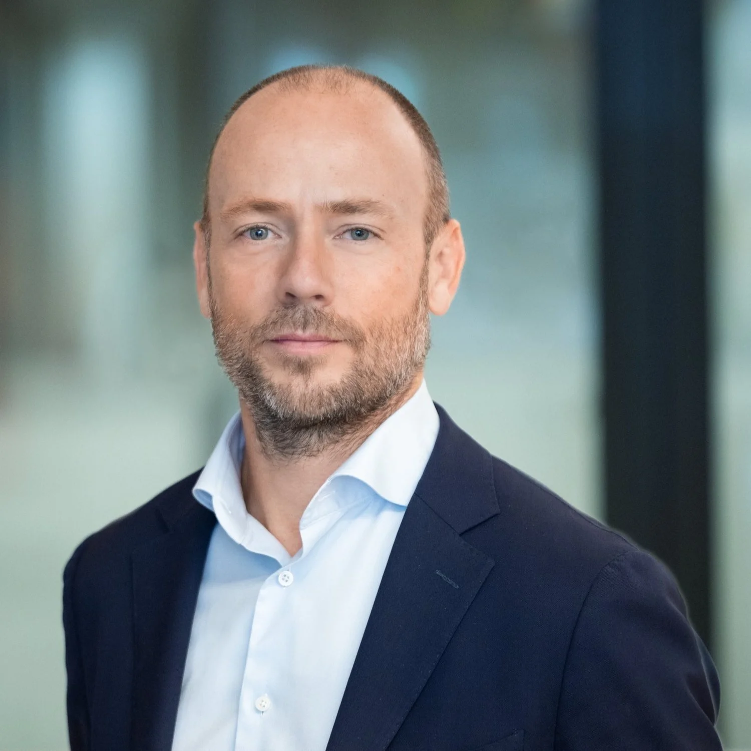 Professional headshot of a middle-aged man with a beard, wearing a dark blue suit and a white dress shirt, in an office setting.