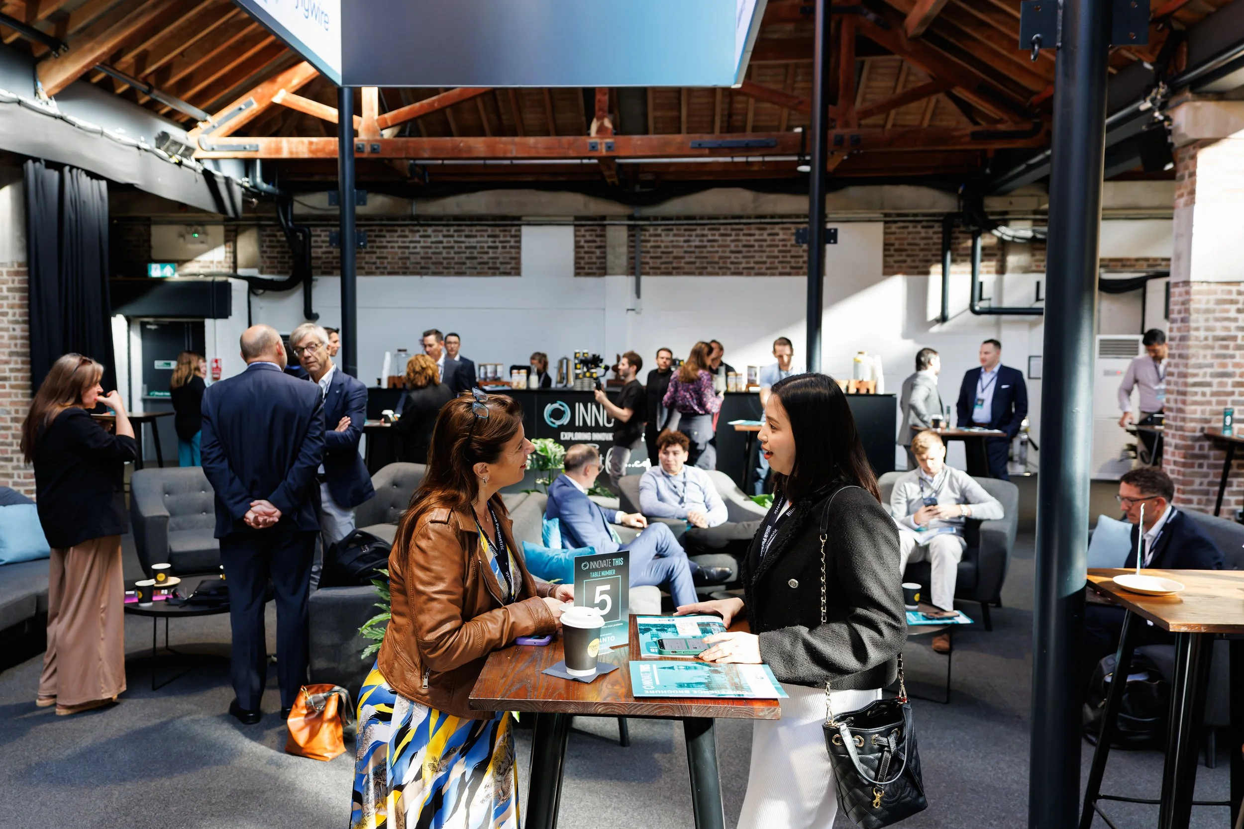 People gathering in a modern, industrial-style conference or event space with exposed brick walls, wooden beams, and black metal supports, engaged in conversations and activities.