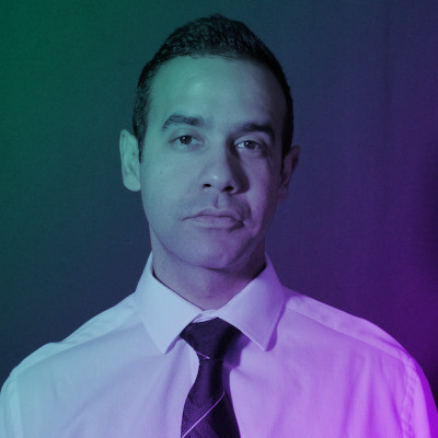 Portrait of a man in a white shirt and dark tie, with colorful lighting effects on a dark background.