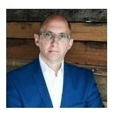 A man wearing glasses and a blue suit jacket, standing indoors in front of a rustic wooden wall.