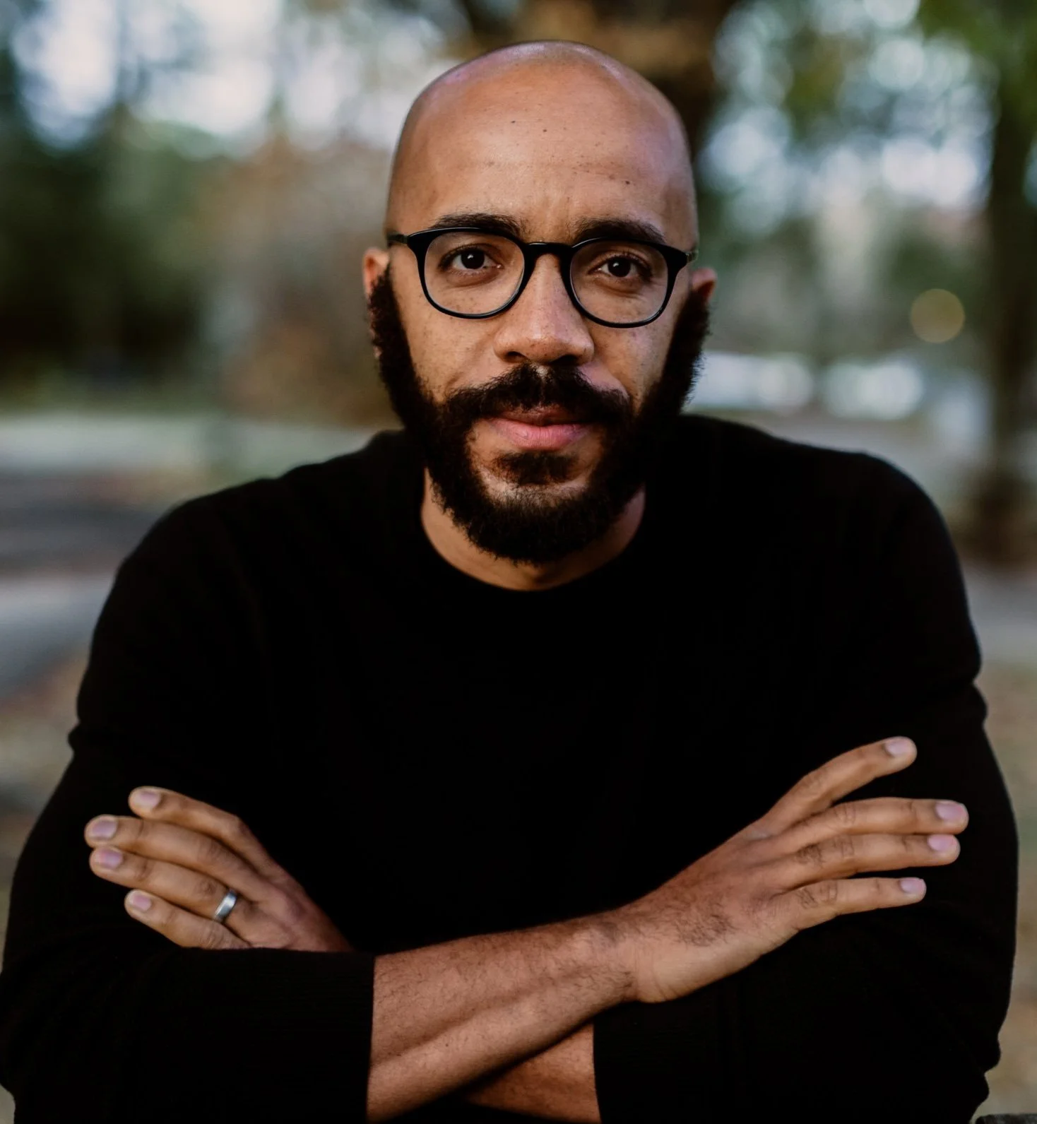 A man with a bald head, full beard, glasses, and a black shirt, sitting outdoors with arms crossed, with blurred trees in the background.