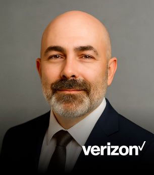 A bald man with a beard wearing a suit and tie, smiling against a gray background with Verizon logo.