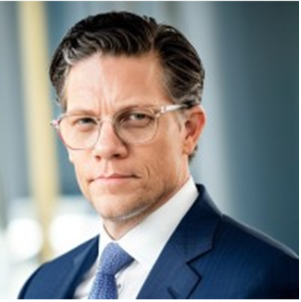 A man in a navy blue suit with a white shirt and light blue tie, wearing glasses, looking serious with furrowed brows.