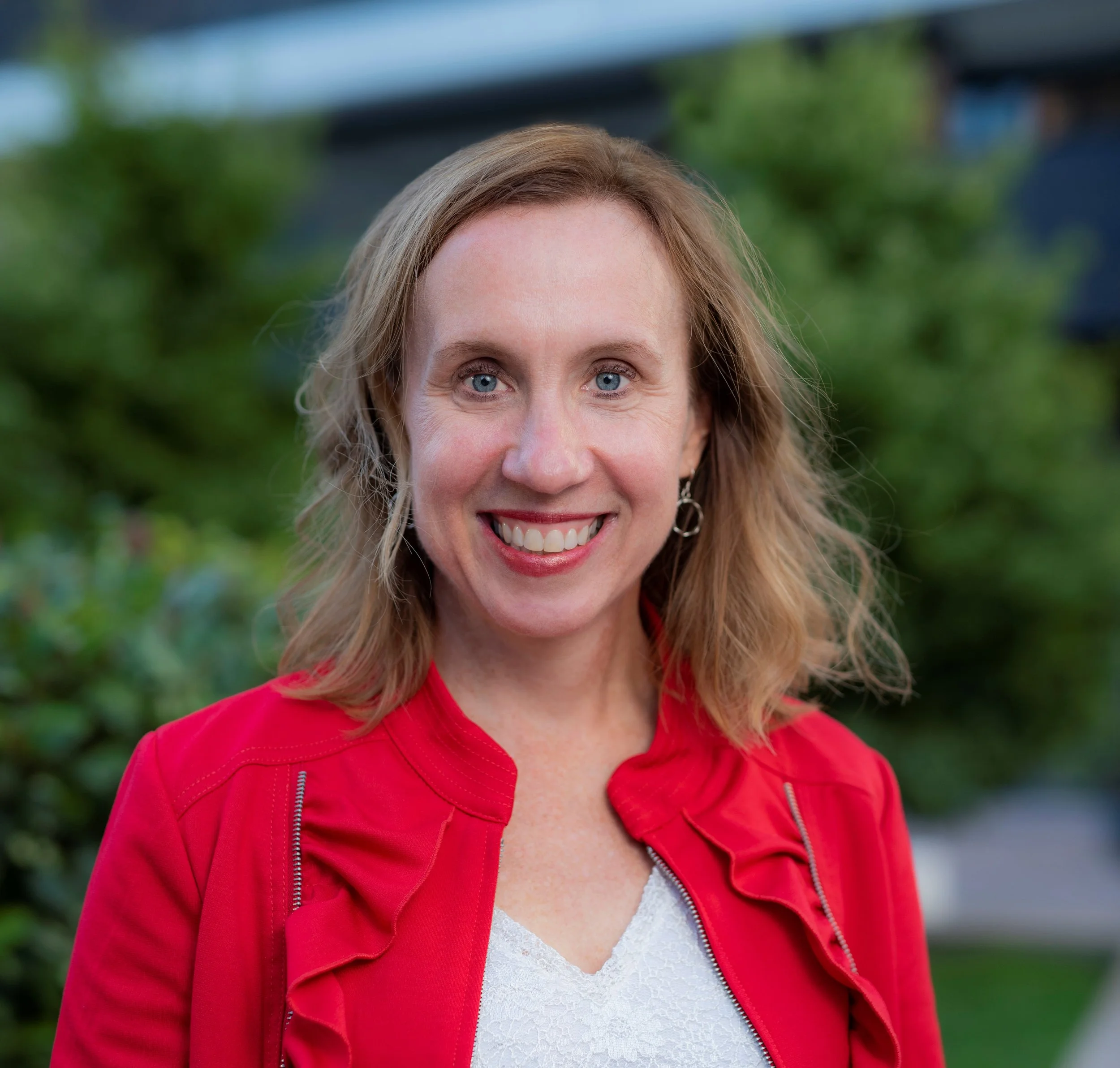 A woman with blonde hair and blue eyes smiling outdoors, wearing a red jacket and earrings, with greenery in the background.