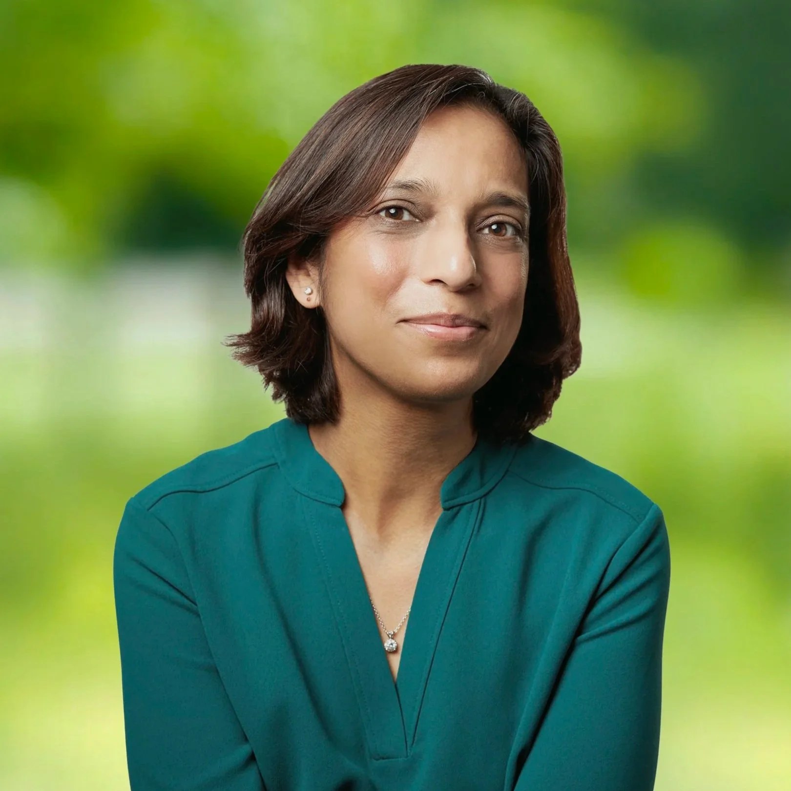 Professional woman with shoulder-length hair wearing a teal top and jewelry, smiling softly against a blurred green outdoor background.