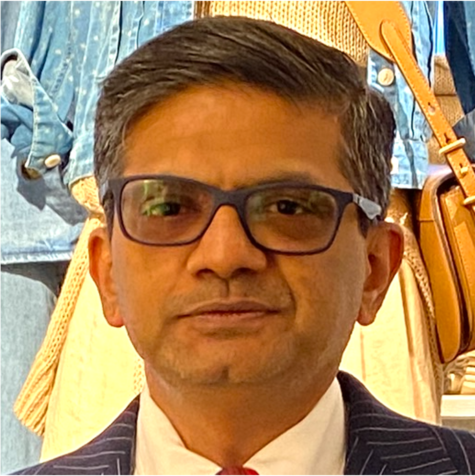 Close-up of a man wearing black glasses, a white shirt, a dark blazer with pinstripes, and a red tie, with a background of blue denim and tan leather bags and accessories.