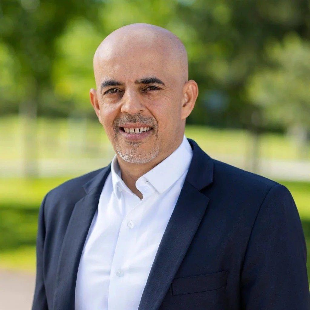 A portrait of a bald man with light skin, wearing a white collared shirt and a dark suit jacket, standing outdoors with a blurred green park background.