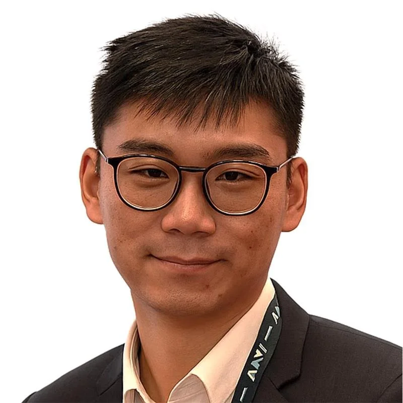 Portrait of a young man wearing glasses in a suit with a lanyard against a white background.