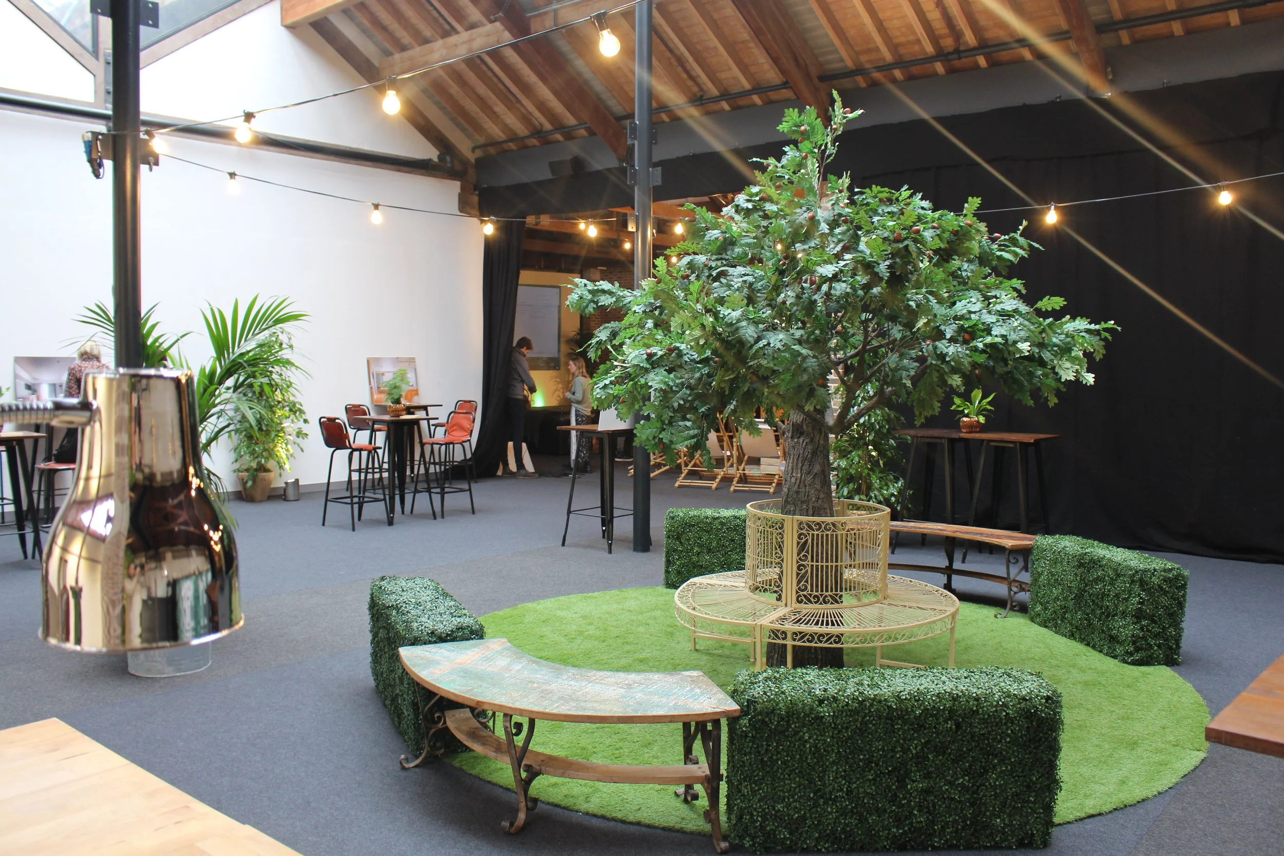 An indoor space with a large potted tree surrounded by a green circular rug and outdoor furniture, including benches and tables. There are string lights hanging from the ceiling, and a few people in the background near a white wall with framed pictures and plants.