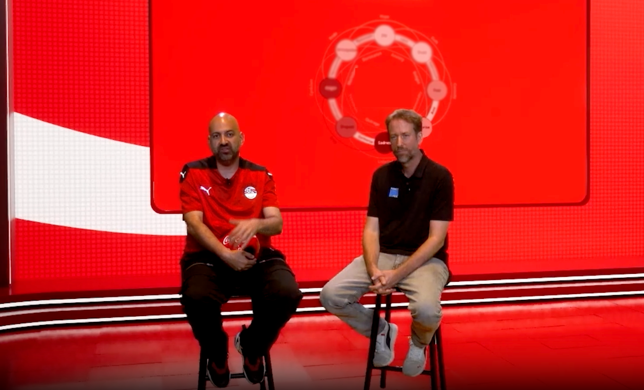 Two men sitting on stools in front of a red background, with a circular diagram on a screen behind them, likely during a sports interview or presentation.