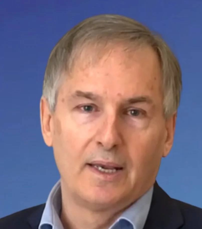 Close-up of a middle-aged man with light-colored hair wearing a dark suit and a light blue shirt, speaking against a plain blue background.