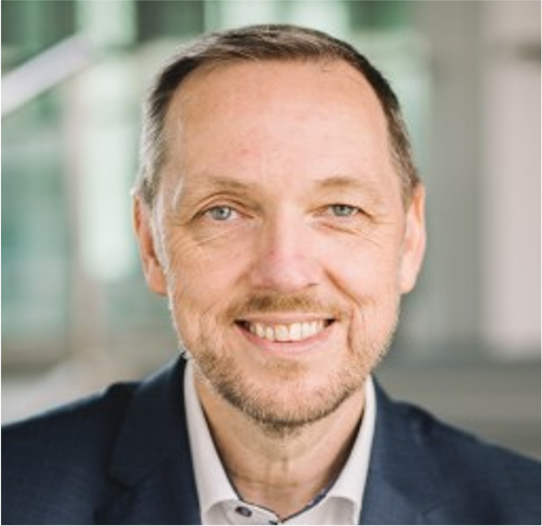 Close-up of a smiling man with short brown hair, blue eyes, and stubble, wearing a suit and white shirt, in an indoor setting with blurred background.