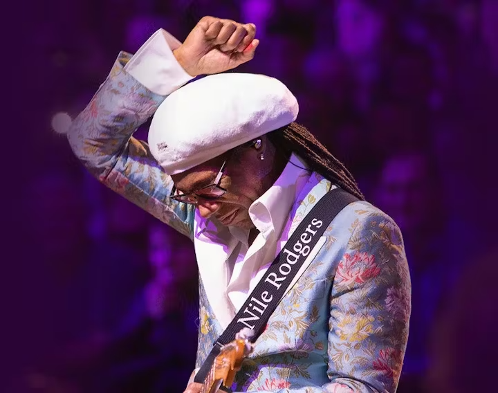 A woman wearing a white beret, glasses, and a floral patterned blazer, playing a guitar, with a strap labeled 'Nile Rodgers'.