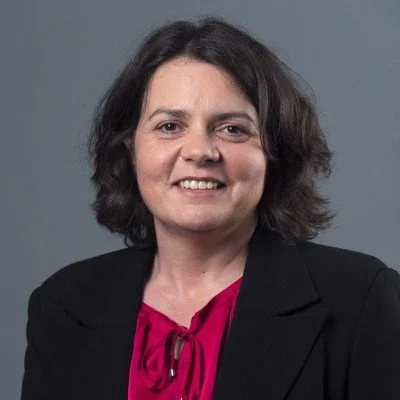 A woman with shoulder-length dark hair, smiling, wearing a black blazer over a pink blouse, against a plain gray background.