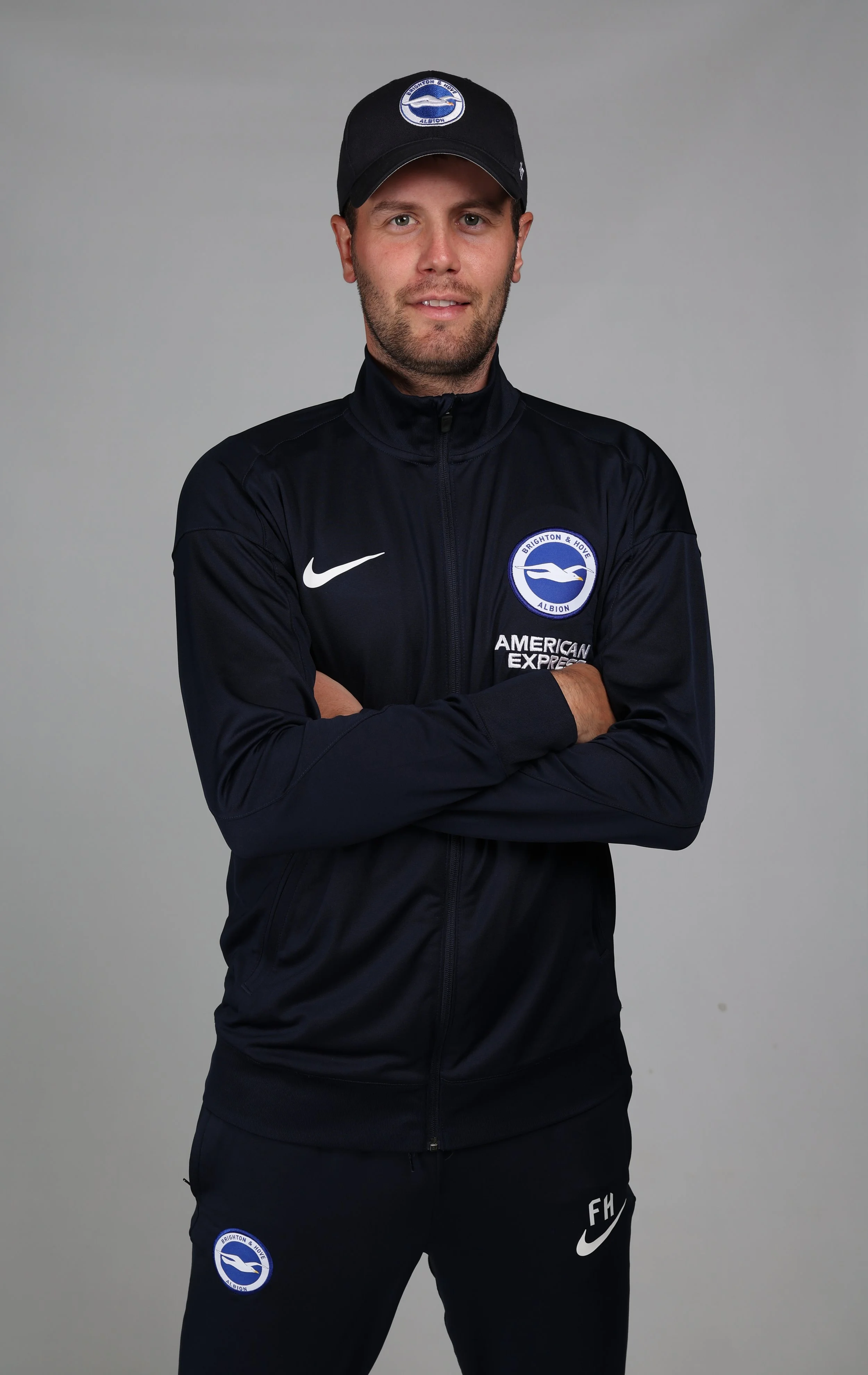 A man standing with arms crossed, wearing a black Brighton & Hove Albion sports jacket and cap, against a plain gray background.