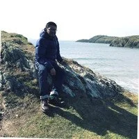 Person standing on rocks near water on a cloudy day, wearing a dark jacket, jeans, and sunglasses.