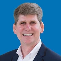 Portrait of a smiling man with short gray hair, wearing a dark suit jacket and a white shirt, set against a blue background.