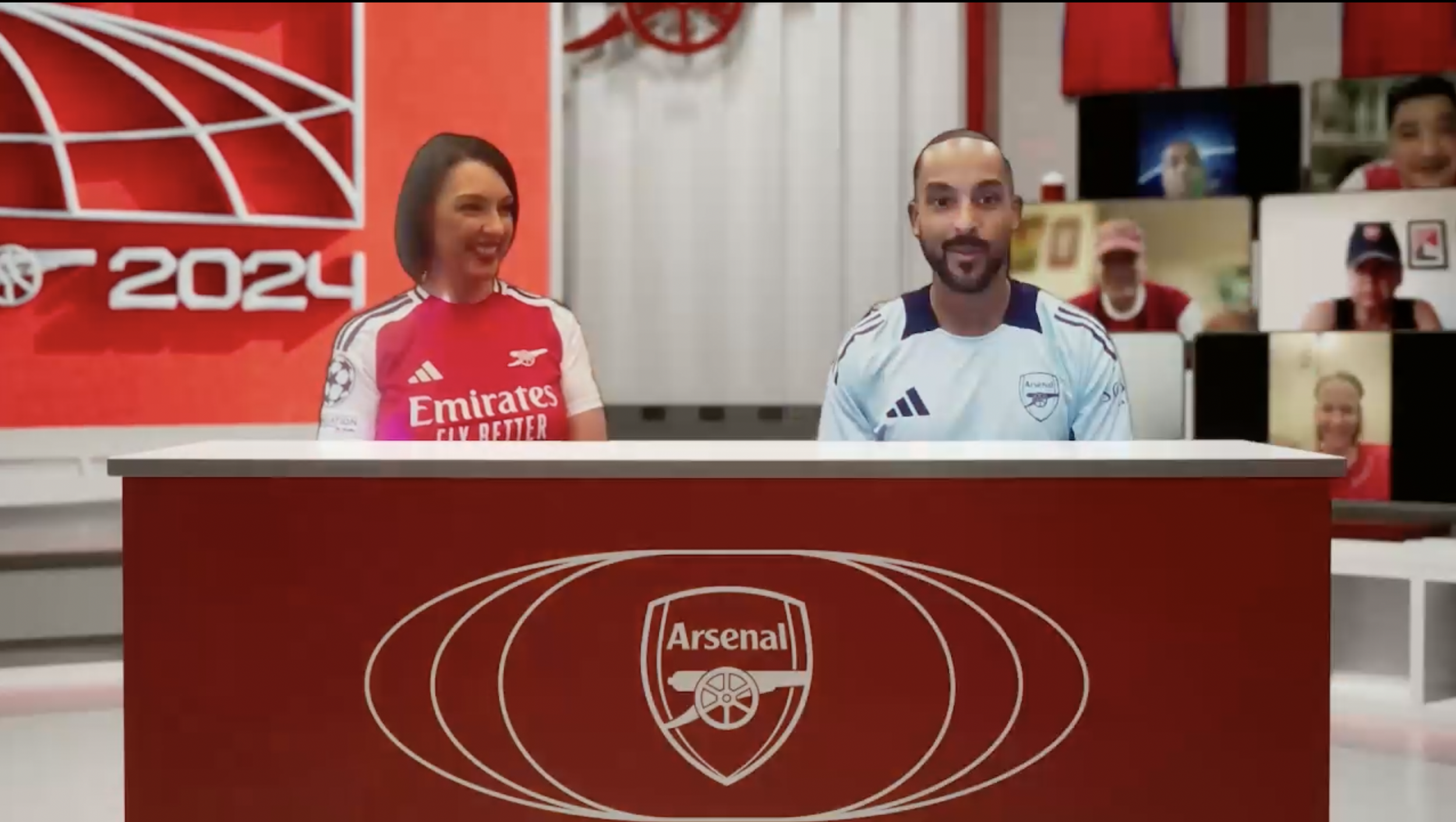 Two people seated at a red desk with an Arsenal logo, participating in a virtual event related to the 2024 season, with multiple screens showing other participants in the background.