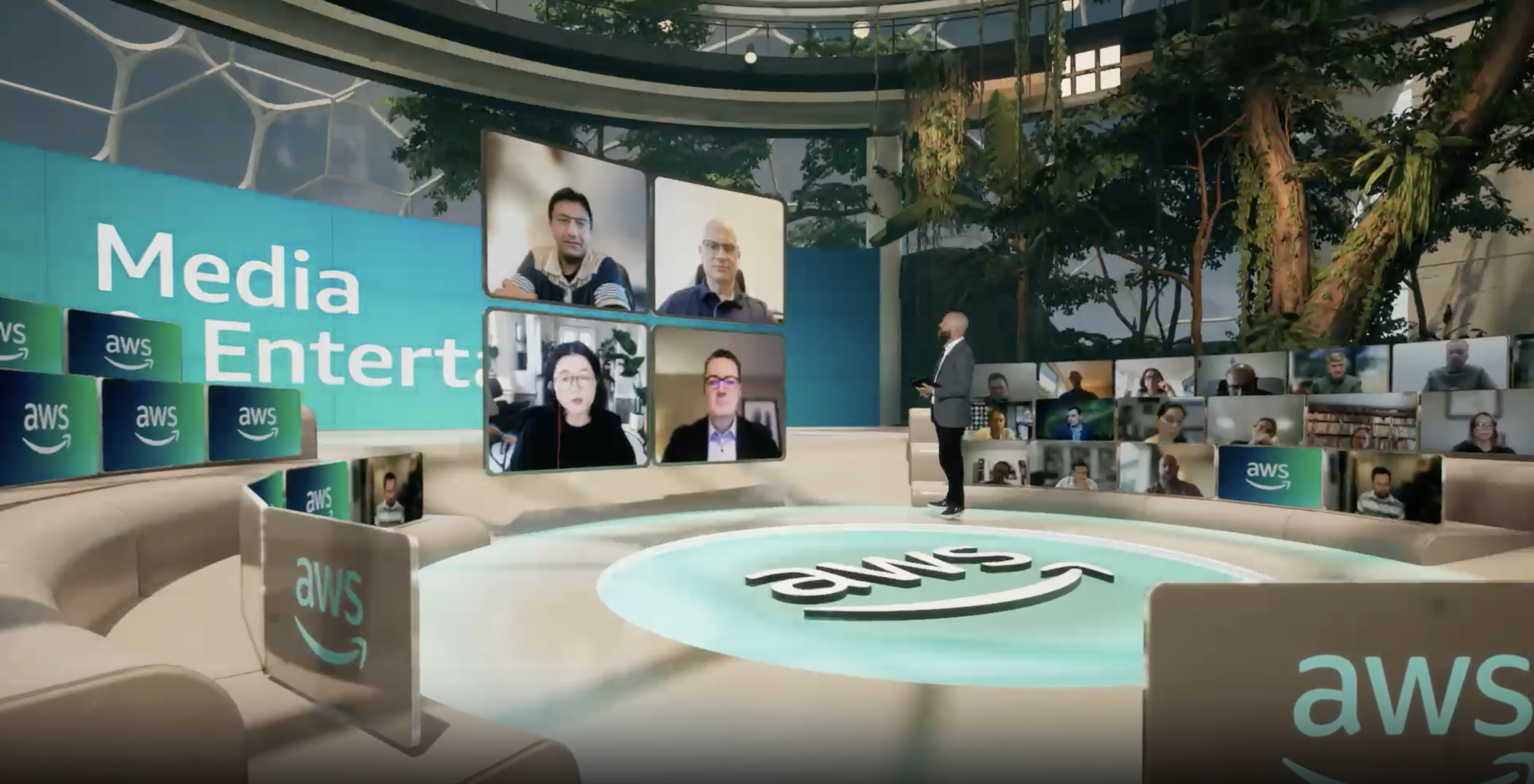 A virtual conference studio with Amazon Web Services branding, featuring a large screen displaying six participants in a video call and multiple smaller screens showing other attendees, with a presenter standing on a circular stage surrounded by Amazon and AWS logos.