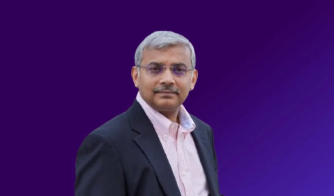 A middle-aged man with gray hair and glasses, wearing a black blazer and a light pink shirt, standing against a plain dark purple background.